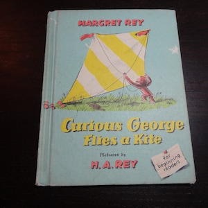 Puede incluir: Una cubierta de libro azul con el título "Curious George Flies a Kite" de Margret Rey con ilustraciones de H.A. Rey. La cubierta presenta una cometa amarilla y blanca con un mono rojo sosteniendo la cuerda.