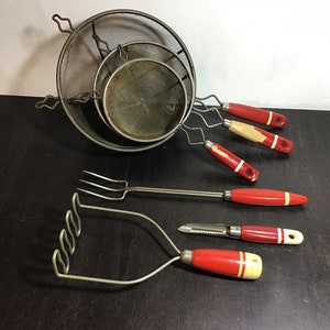 May include: Set of vintage kitchen tools with red and white handles. The set includes a potato masher, a fork, a peeler, and three wire mesh strainers.