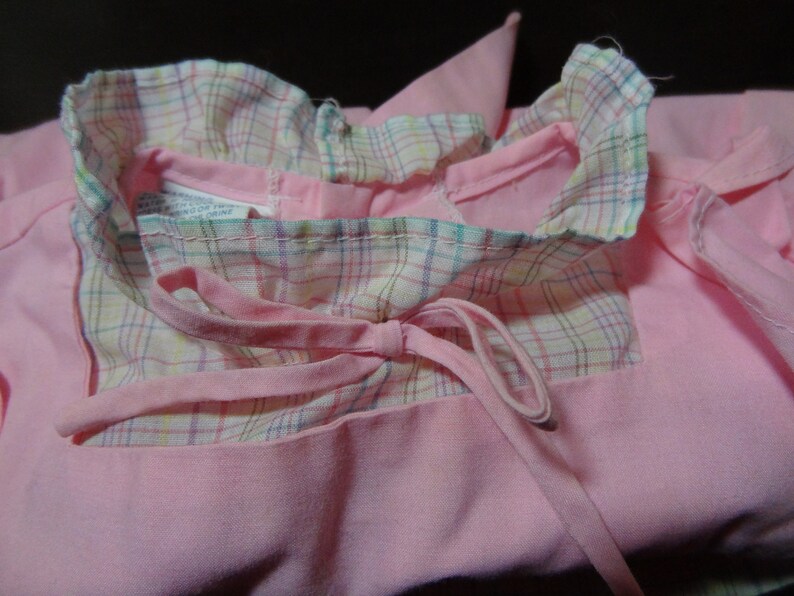 May include: A pink dress with a plaid collar and a bow tie. The dress has a ruffled collar and a tie that can be tied in the front.