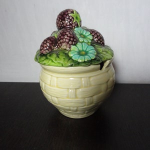 May include: A ceramic sugar bowl with a lid shaped like a basket overflowing with purple and green berries and flowers. The bowl is yellow with a woven basket pattern.