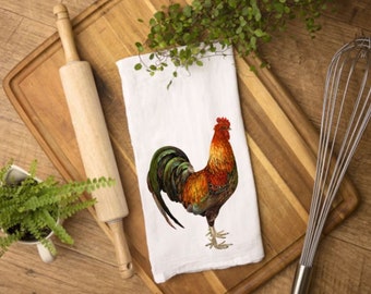 May include: A white kitchen towel with a colorful rooster illustration. The rooster has red, orange, green, and brown feathers. A wooden rolling pin, cutting board, and whisk are also in the image, suggesting a kitchen theme.