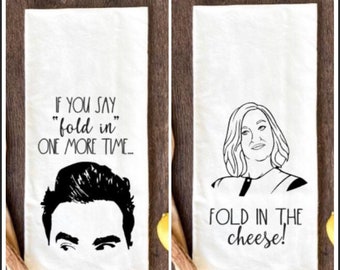May include: Two white kitchen towels with black text and illustrations. One towel says "IF YOU SAY 'fold in' ONE MORE TIME..." with a black illustration of a man's face. The other says "FOLD IN THE cheese!" with a woman's face.