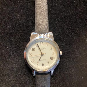 May include: A silver-toned quartz watch with a white face and black leather band. The watch face features black numerals, a small heart, and the word "MEURIO". The watch has a bow-shaped design at the top.