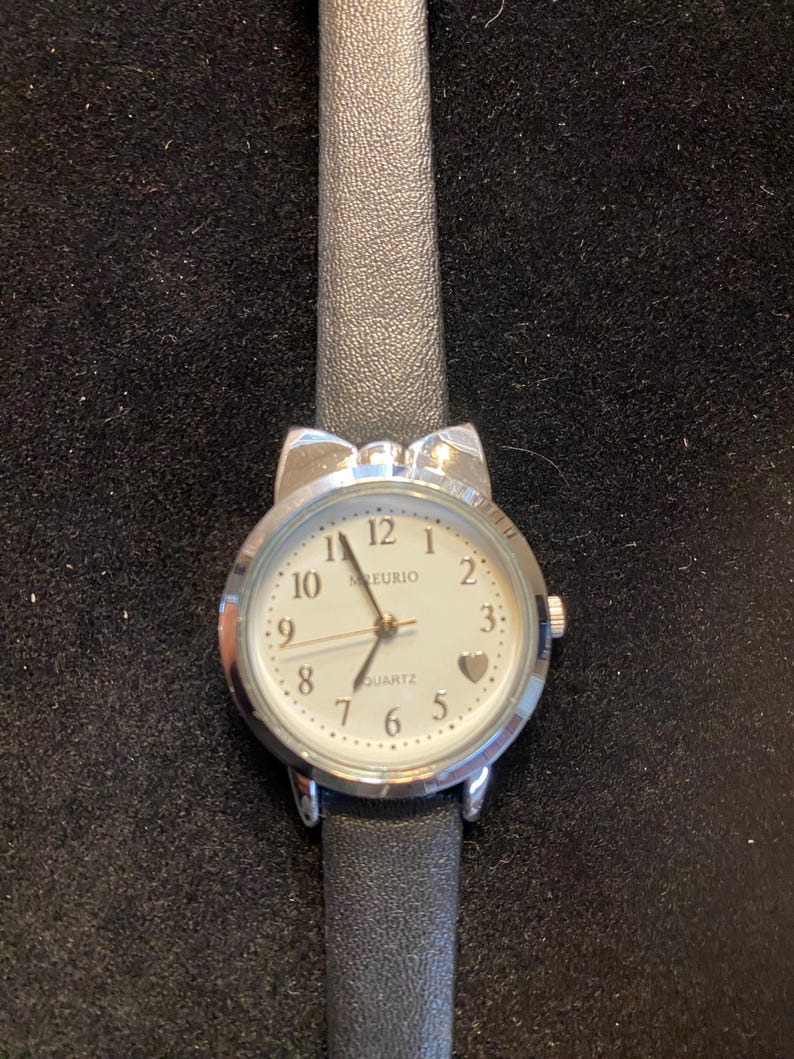 May include: A silver-toned quartz watch with a white face and black leather band. The watch face features black numerals, a small heart, and the word "MEURIO". The watch has a bow-shaped design at the top.