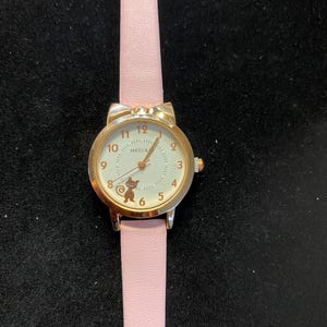 May include: A gold-toned wristwatch with a light pink band. The watch face is white with black numerals and a small cat illustration. The band is light pink and made of a smooth material. The watch has a bow-shaped top.
