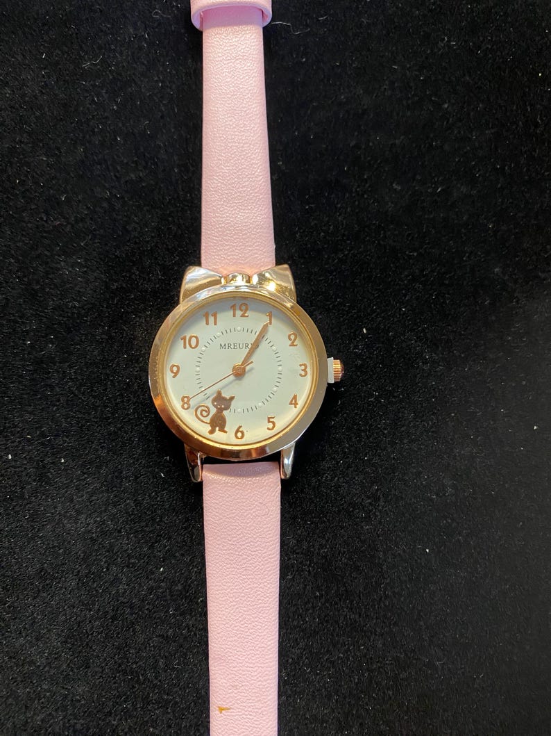 May include: A gold-toned wristwatch with a light pink band. The watch face is white with black numerals and a small cat illustration. The band is light pink and made of a smooth material. The watch has a bow-shaped top.