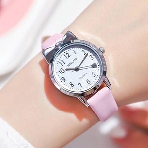May include: A pink wristwatch with a silver bow-shaped bezel. The watch face is white with black numerals and hands. The watch band is light pink.