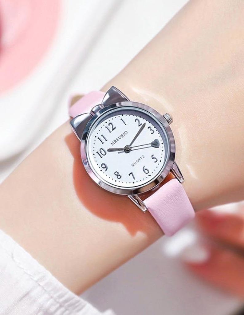May include: A pink wristwatch with a silver bow-shaped bezel. The watch face is white with black numerals and hands. The watch band is light pink.