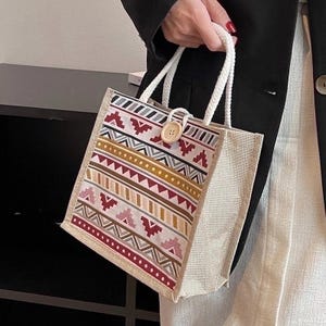 May include: A small tote bag with a geometric pattern in shades of red, pink, yellow, and brown. The bag has a natural-colored burlap body, white rope handles, and a wooden button closure. The bag is held by a person.