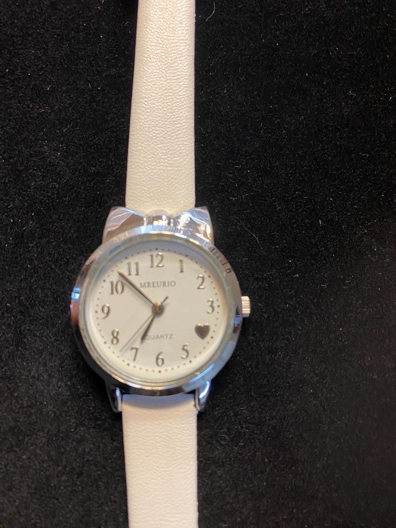 May include: A silver-toned quartz watch with a white face and a white leather band. The watch face features black numerals, a heart-shaped marker, and the brand name "MREURIO". The band is approximately 0.5 inches wide.