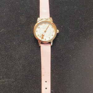 May include: A gold-toned wristwatch with a light pink leather band. The watch face has a white background with black numerals and a small cat silhouette. The band has a buckle closure and multiple adjustment holes.