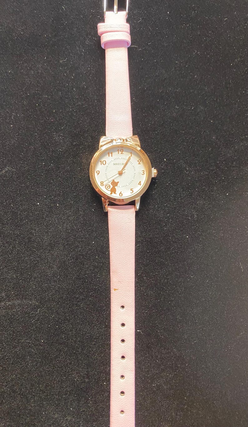 May include: A gold-toned wristwatch with a light pink leather band. The watch face has a white background with black numerals and a small cat silhouette. The band has a buckle closure and multiple adjustment holes.