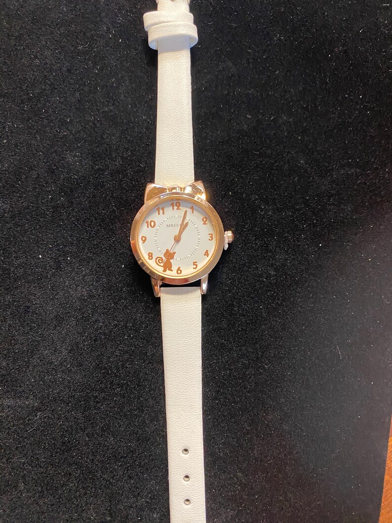 May include: A white wristwatch with a gold-colored cat ear-shaped bezel. The watch face is white with gold-colored numerals and hands. The watch has a white leather strap.