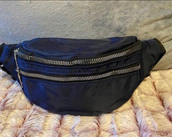 Può includere: Marsupio blu navy con due scomparti con cerniera e hardware color argento. La borsa è realizzata in un materiale liscio. Il marsupio è mostrato su una superficie rosa e bianca strutturata.