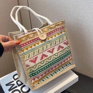 May include: A square tote bag with a beige woven body and a colorful patterned front panel. The panel features geometric designs in red, green, yellow, and pink. It has white rope handles and a large wooden button closure.