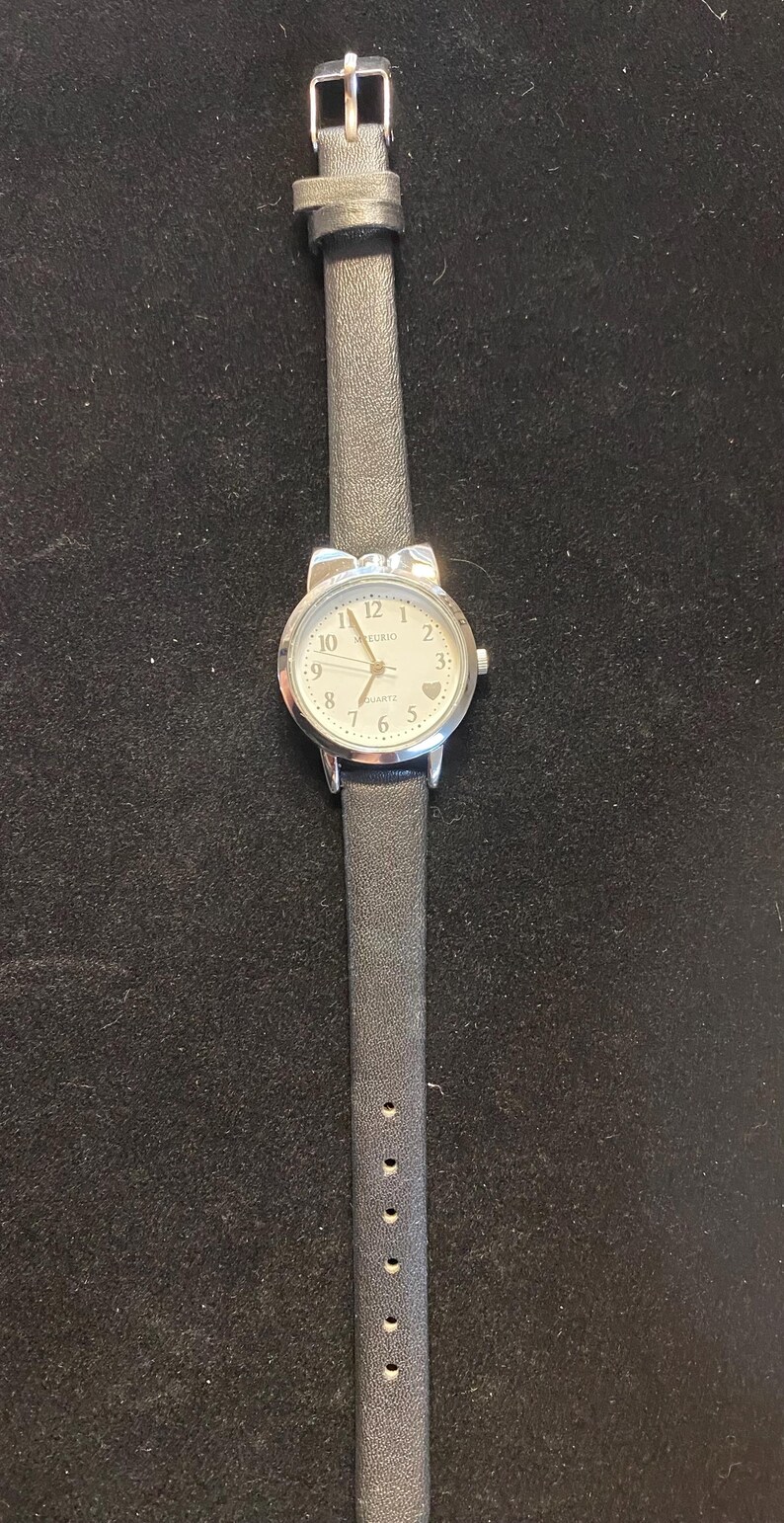 May include: A silver-toned analog watch with a white face and black leather band. The watch face features black numerals and hands, with the brand name "Kurio" at the top. The band is approximately 0.5 inches wide.