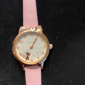 May include: A gold-toned wristwatch with a pink leather band. The watch face is white with black numerals and a small cat silhouette. The watch case has cat ear-shaped accents. The watch hands are gold-toned.