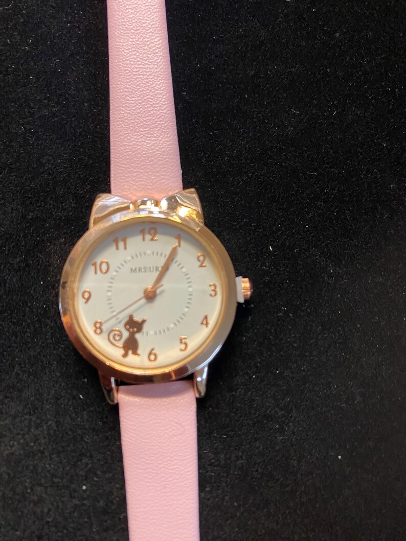 May include: A gold-toned wristwatch with a pink leather band. The watch face is white with black numerals and a small cat silhouette. The watch case has cat ear-shaped accents. The watch hands are gold-toned.