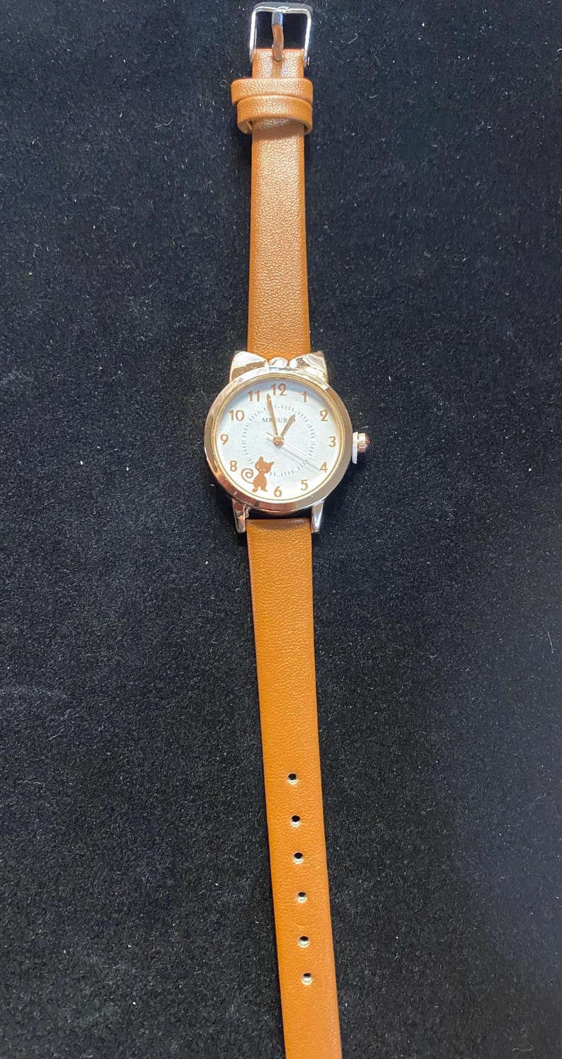 May include: A gold-toned wristwatch with a white face and brown leather band. The watch face features black numerals and a small cartoon character. The band is a light brown color and has a buckle closure.