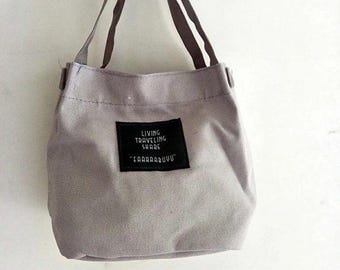 May include: A small, pale grey tote bag with two brown handles. A black rectangular patch on the front displays the text "LIVING TRAVELING SHARE" and "TAAHAADUVU" in white. The bag is held up by a hand.