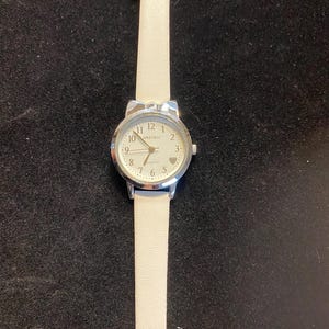May include: A silver-toned wristwatch with a white leather band. The watch face is white with black numerals and hands, and the brand name "MBREURIO" is printed on the face. A small heart is at the 3 o'clock position.