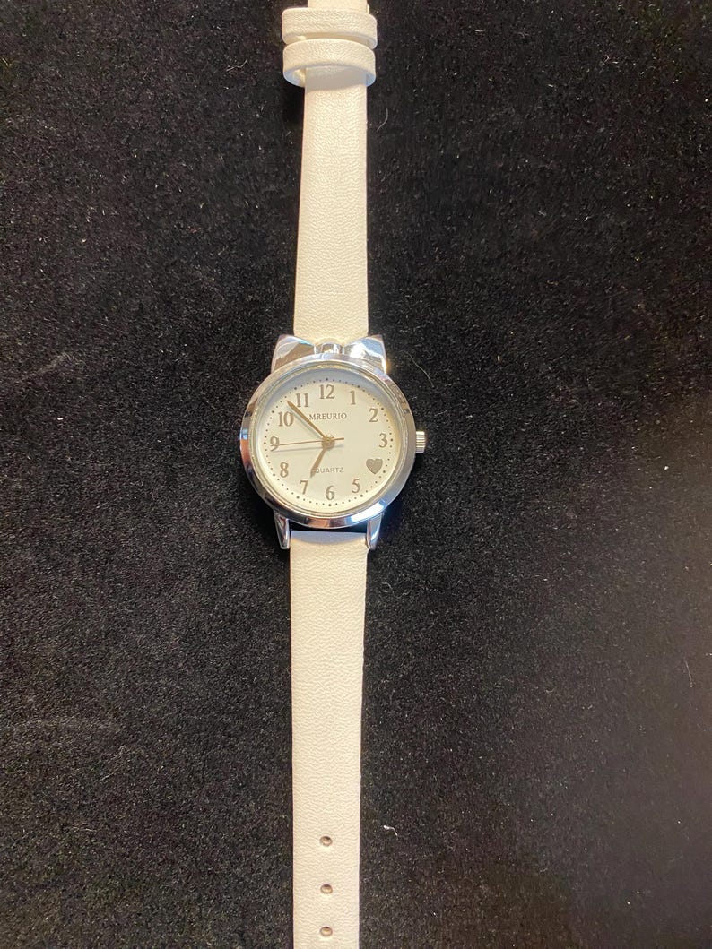 May include: A silver-toned wristwatch with a white leather band. The watch face is white with black numerals and hands, and the brand name "MBREURIO" is printed on the face. A small heart is at the 3 o'clock position.