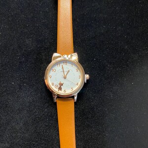 May include: A silver-toned wristwatch with a brown leather band. The watch face is white with gold-colored numerals and a small cat silhouette. The watch has a cat ear design at the top of the watch face.