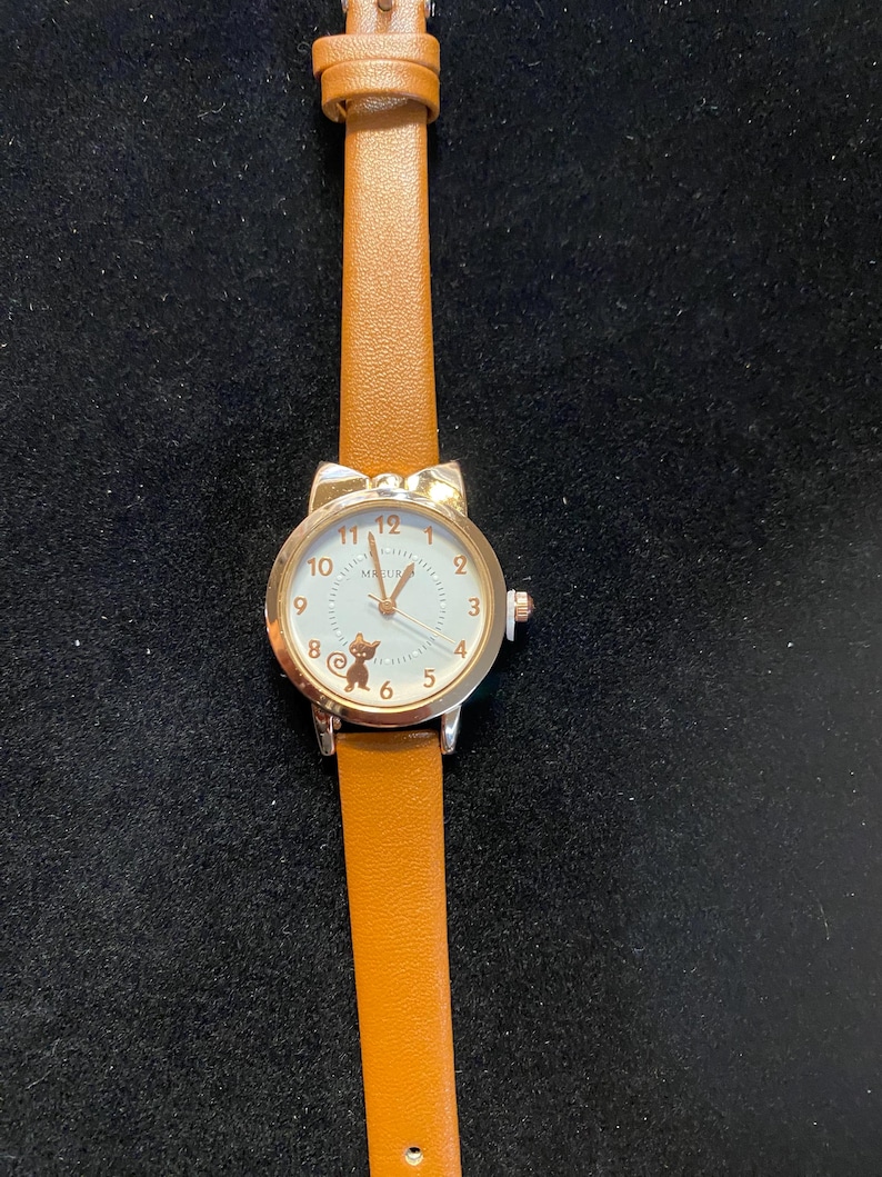 May include: A silver-toned wristwatch with a brown leather band. The watch face is white with gold-colored numerals and a small cat silhouette. The watch has a cat ear design at the top of the watch face.