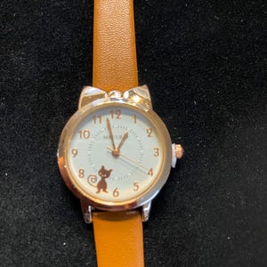 May include: A silver-toned wristwatch with a tan leather band. The watch face is white with black numerals and a small cat graphic. The watch case has cat ear accents. The watch reads "MEUR".