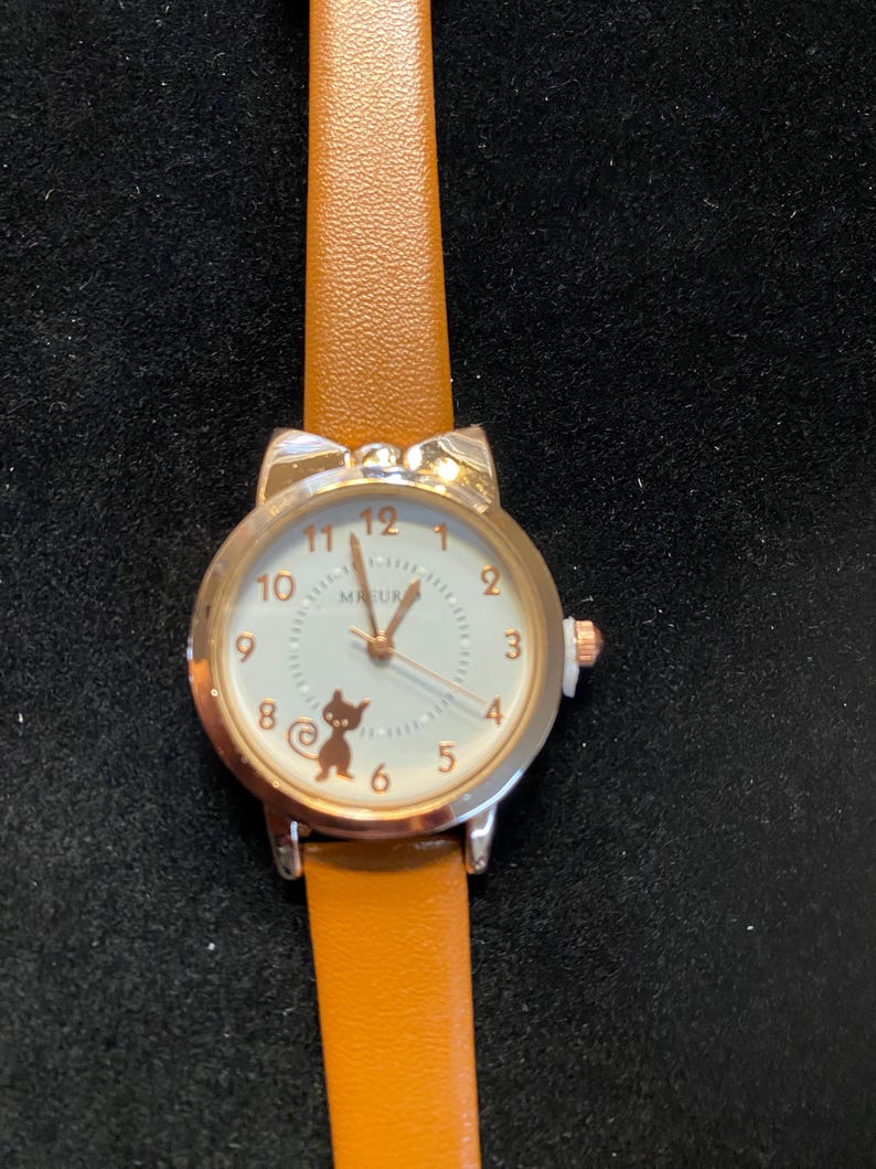 May include: A silver-toned wristwatch with a tan leather band. The watch face is white with black numerals and a small cat graphic. The watch case has cat ear accents. The watch reads "MEUR".