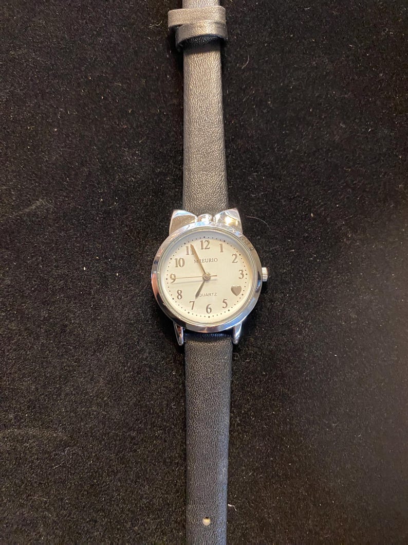 May include: A silver-toned wristwatch with a white face and black leather band. The watch face has black numerals and a small heart at the 3 o'clock position. The band is approximately 0.5 inches wide.