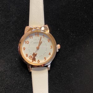 May include: A gold-toned wristwatch with a white face and a white leather strap. The watch face features gold-colored numerals and a small bear graphic. The watch has a round face with a cat ear design at the top.