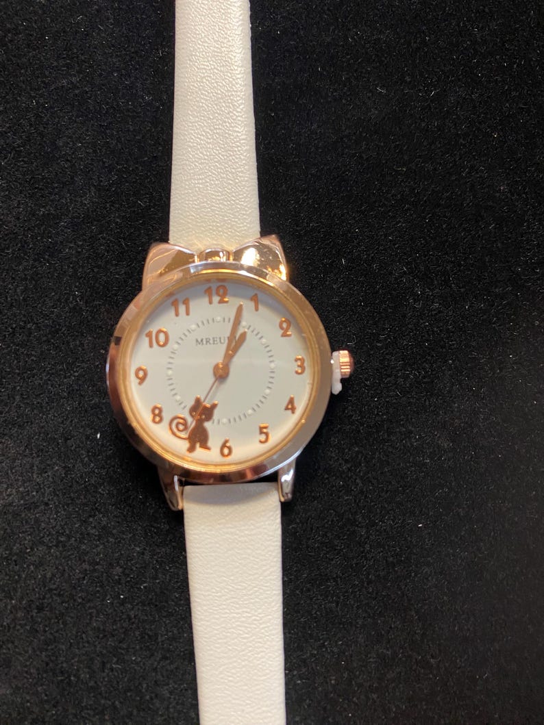 May include: A gold-toned wristwatch with a white face and a white leather strap. The watch face features gold-colored numerals and a small bear graphic. The watch has a round face with a cat ear design at the top.