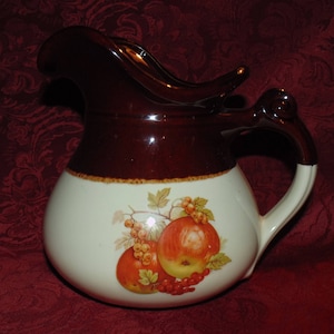 Könnte beinhalten: Eine braune und weiße Keramikkanne mit Henkel und Ausguss. Die Kanne hat ein Design mit Äpfeln und Beeren auf der Vorderseite.