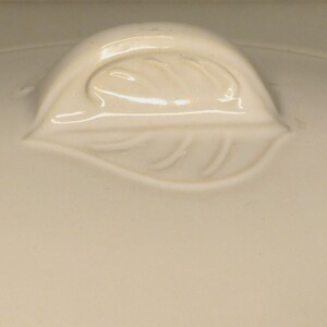 Puede incluir: Tapa de cer&aacute;mica color crema con un asa en forma de hoja. El asa est&aacute; en relieve y detallada con vetas de hojas. La tapa tiene un acabado liso y brillante, lo que sugiere un art&iacute;culo de cocina o comedor.