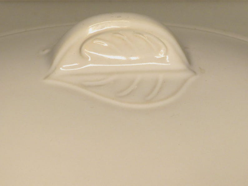 Puede incluir: Tapa de cer&aacute;mica color crema con un asa en forma de hoja. El asa est&aacute; en relieve y detallada con vetas de hojas. La tapa tiene un acabado liso y brillante, lo que sugiere un art&iacute;culo de cocina o comedor.