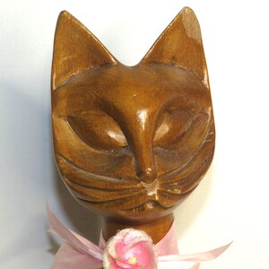 Puede incluir: Una figurita de gato de madera con un lazo rosa y una flor rosa. El gato tiene una cara tallada de color marr&oacute;n con los ojos cerrados.