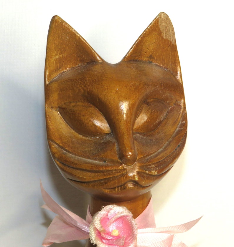 Puede incluir: Una figurita de gato de madera con un lazo rosa y una flor rosa. El gato tiene una cara tallada de color marr&oacute;n con los ojos cerrados.