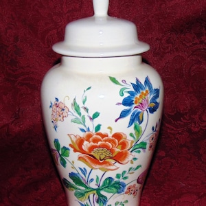 May include: A white ceramic jar with a lid, decorated with a floral pattern in shades of blue, orange, green, and pink. The jar has a classic, elegant design.