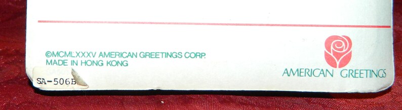 May include: White rectangular card with the text "&copy;MCMLXXXV AMERICAN GREETINGS CORP. MADE IN HONG KONG" and a red rose logo. The card has a torn corner with the text "SA-506B".