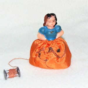 May include: A vintage porcelain doll pincushion with a blue dress and orange skirt. The doll is holding a spool of thread.