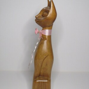 Puede incluir: Una figurita de gato de madera con una cinta rosa alrededor del cuello. El gato est&aacute; de pie sobre una base de madera.
