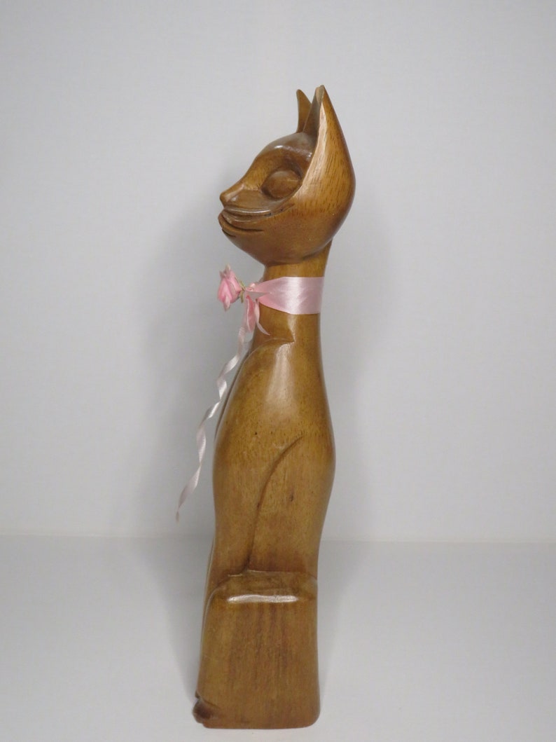 Puede incluir: Una figurita de gato de madera con una cinta rosa alrededor del cuello. El gato est&aacute; de pie sobre una base de madera.