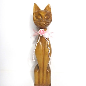 Puede incluir: Una figurita de gato de madera con un lazo rosa y una flor rosa. El gato est&aacute; sentado derecho con los ojos cerrados.