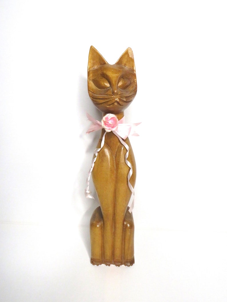 Puede incluir: Una figurita de gato de madera con un lazo rosa y una flor rosa. El gato est&aacute; sentado derecho con los ojos cerrados.