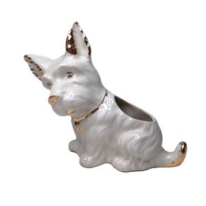 Könnte beinhalten: Ein weißer Keramik-Pflanztopf in Hundeform mit goldenen Akzenten an Ohren, Nase und Schwanz. Der Pflanztopf hat eine kleine Öffnung für Pflanzen. Der Hund sitzt.