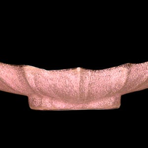 May include: A pink, scalloped-edge bowl with a textured surface. The bowl has a low, rectangular base and a wide, curved shape. The rim has a darker, bronze-colored edge.