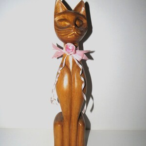 Puede incluir: Una figurita de gato de madera con un lazo rosa y una flor rosa. El gato est&aacute; sentado derecho con los ojos cerrados.