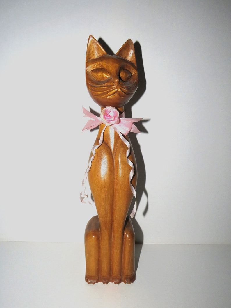 Puede incluir: Una figurita de gato de madera con un lazo rosa y una flor rosa. El gato est&aacute; sentado derecho con los ojos cerrados.