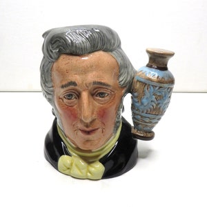 May include: A ceramic mug shaped like a man's head with a gray wig and a blue and brown vase handle. The mug is painted with realistic details and features a black jacket and a white cravat.